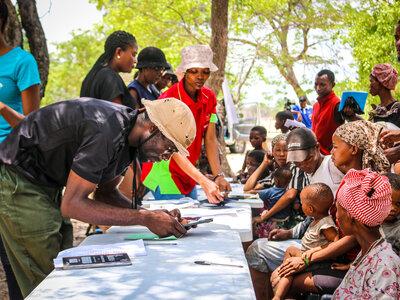 Photo: WFP/Theodor Uukongo People gather around a table outdoors as staff register households using mobile devices and paper forms, while caregivers sit with children nearby under trees.