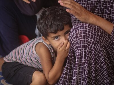Child leans on adult in patterned garment; adult's hand rests gently on child's head in a comforting gesture.