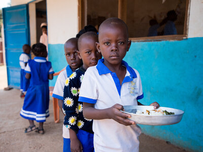 Photo: WFP/Aurore Vinot Children in school uniforms lined up outside blue-walled building; one child holds a plate of rice and vegetables, others wait in background.