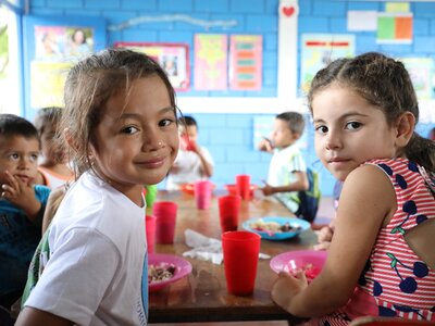 Children sitting around a table in a colourful classroom, eating meals served in bright bowls with red cups.