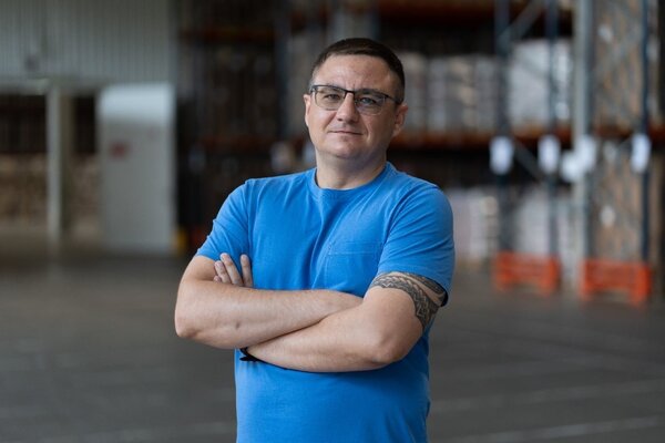 Person stands in a warehouse with arms crossed, wearing a blue shirt, surrounded by stacked pallets and storage racks.