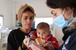 A baby looks into camera, held by her mother, as  nutritionist looks on wearing a facemask