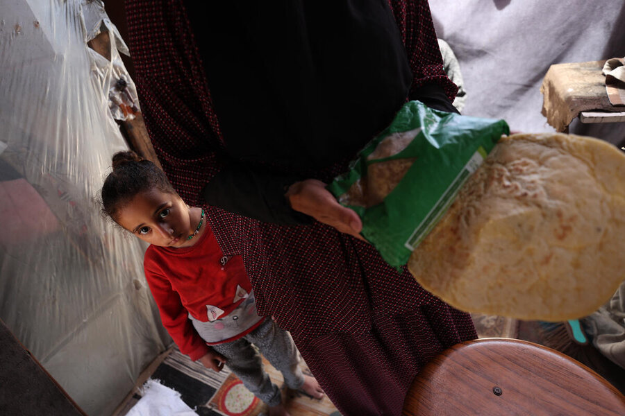 A girl looks into her camera from behind her mother who is holding up a flatbread and a pack pasta