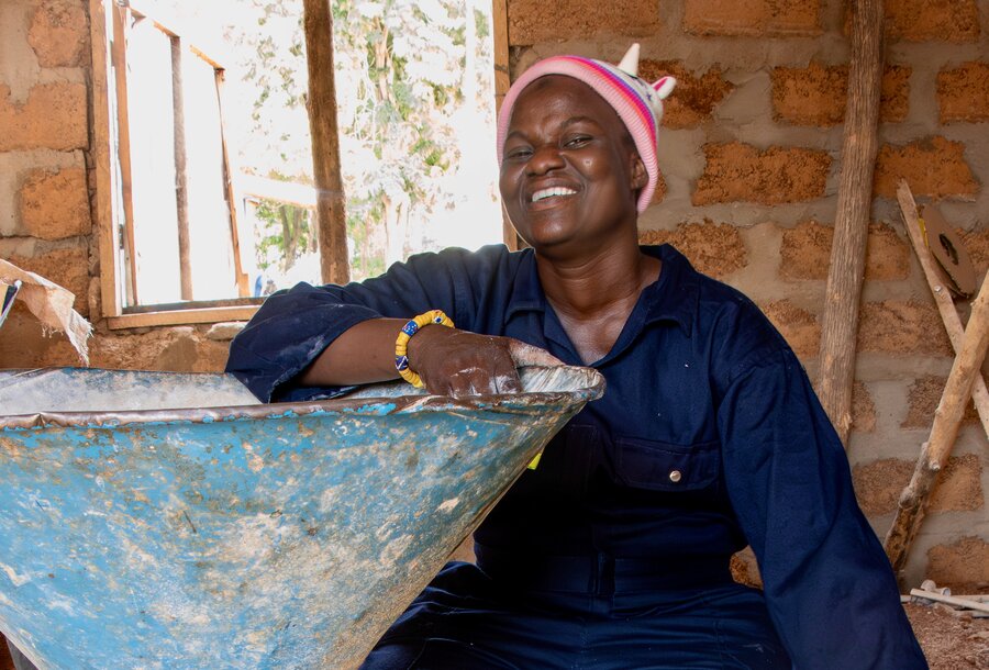 A smiling woman in a red cap and dark blue overalls sits next to a metal container. Photo: WFP/Abdul-Wahab