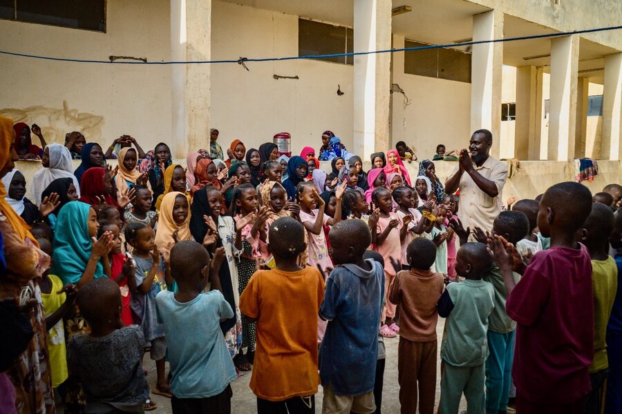 Children in colourful T-shirts gather in a circle singing, with a man in a white shirt leading them. Photo: WFP/Abubakar Garlnabei