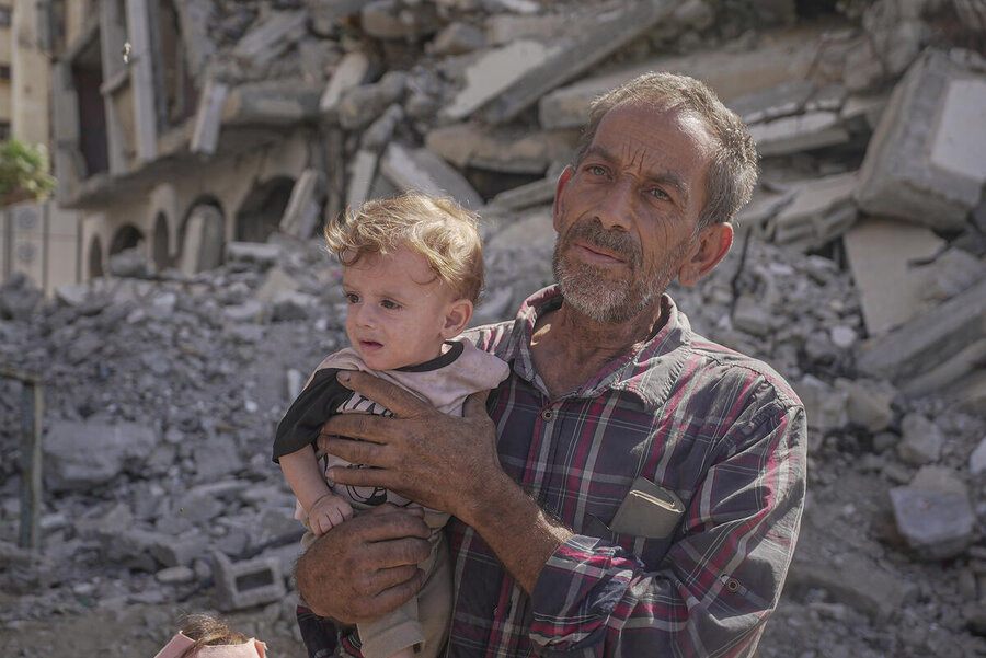 A late middle aged man holds up a small child against a background of rubble in Gaza