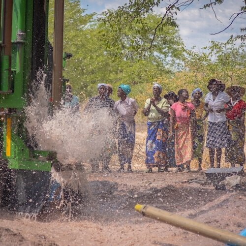 Water gushes from a drilling machine as a group of people stand nearby observing in a rural area.