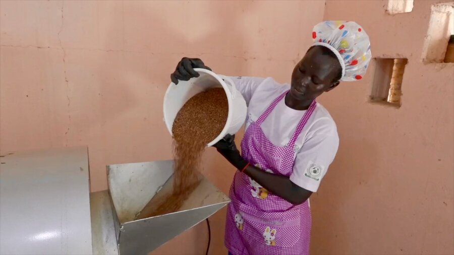 A woman in a white T-shirt and cap, and lavender apron pours grains into a processing machine. Photo: WFP