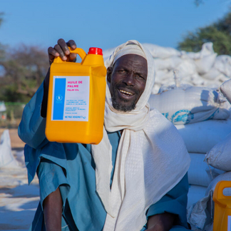 A smiling man holds up a container of palm oil distributed as part of a humanitarian food assistance programme, with stacked relief sacks visible in the background.