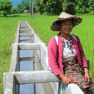 Community members work together to clear and level a hillside path using hand tools. Several people stand and dig along the earthen track, surrounded by trees and vegetation, with forested mountains stretching across the background.
