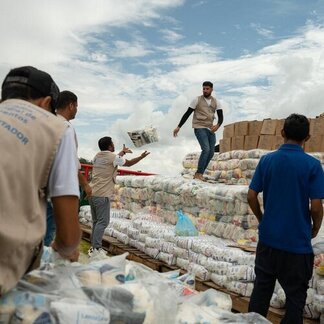 Workers unload and stack food supplies from a boat, tossing packages onto large piles of bags and boxes under a cloudy sky during a relief operation.
